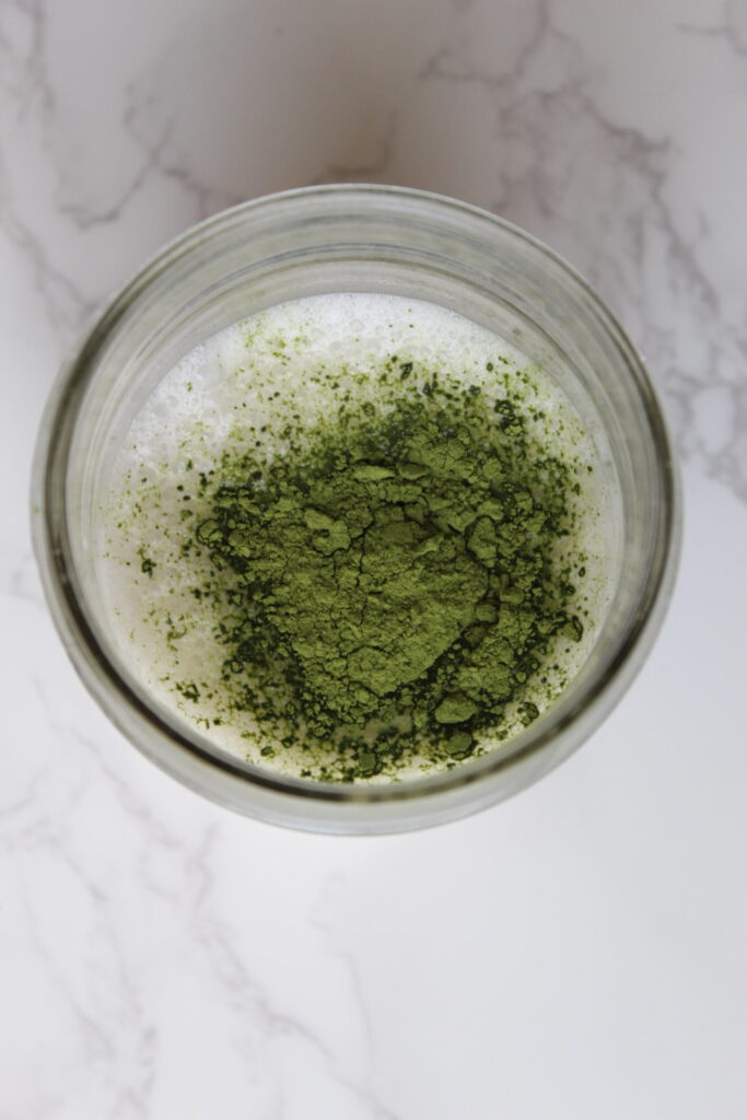 the top view of a mason jar glass filled with almond milk and vibrant green matcha powder sitting on top of the milk. The cup is on a marble countertop.