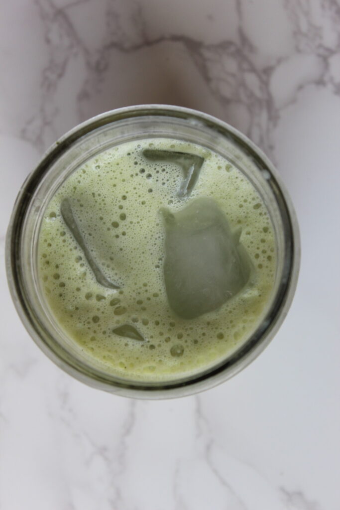 the top view of a mason jar glass with a matcha latte in it and a few ice cubes, sitting on a marble counter top.
