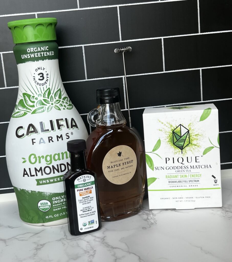 A bottle of almond milk, a container of vanilla extract, a box of matcha tea and a glass bottle of maple syrup all sitting on a kitchen counter with black subway tile in the background.
