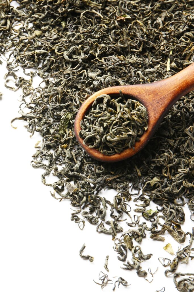 Dried green tea leaves in a spoon and lying all around it on a white countertop