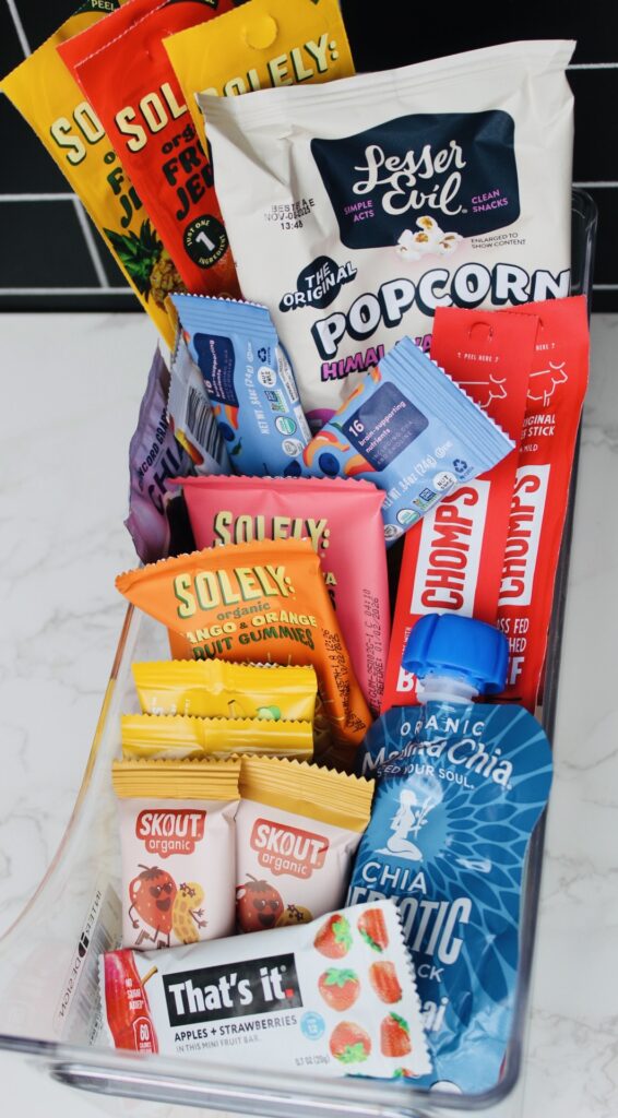 a snack container on a marble counter holding many different toddler and kids snacks such as fruit strips, popcorn, beef sticks, granola bars. 