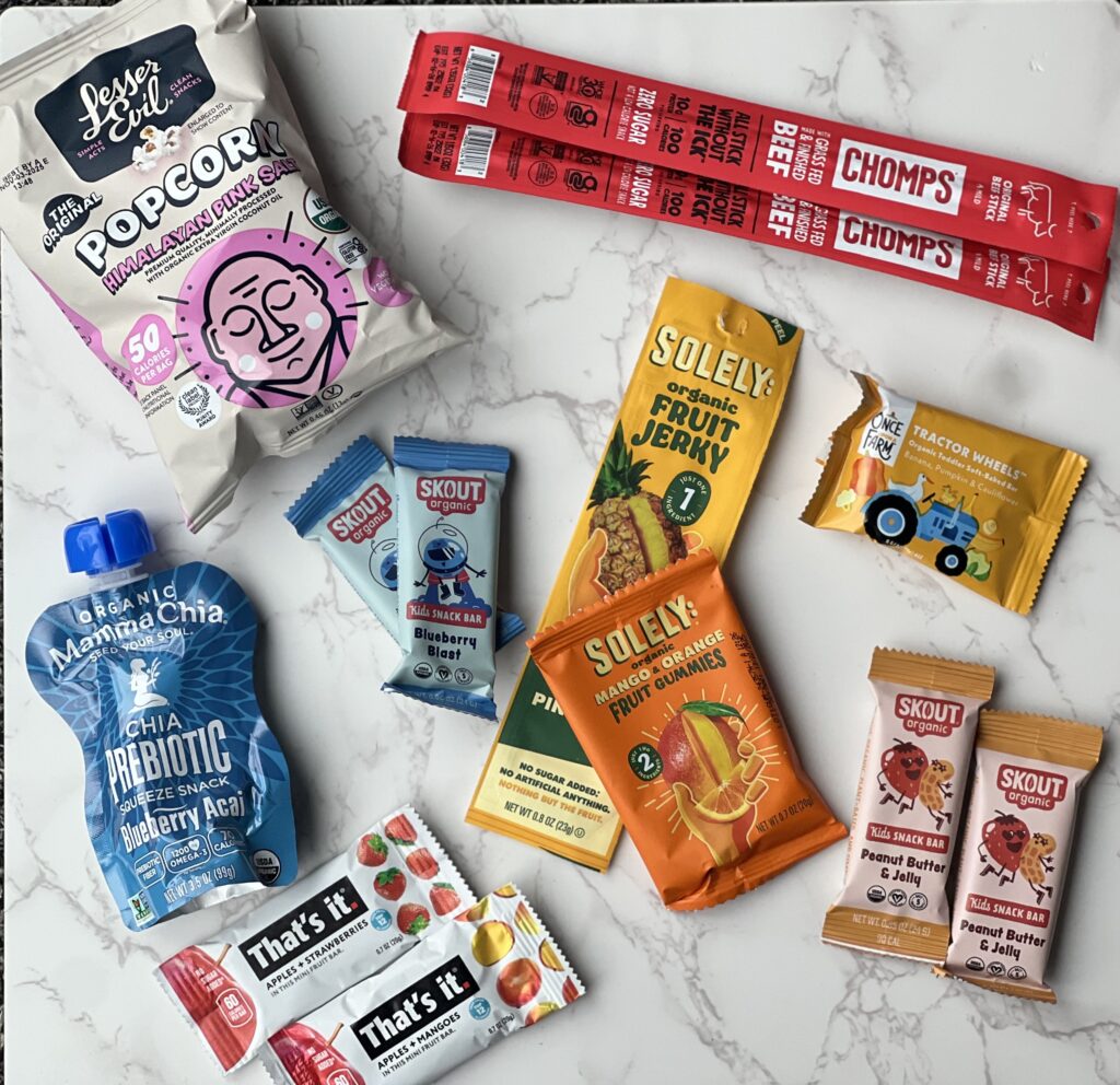 There are some minimally processed snacks lying on a marble countertop, gummies, fruit bars, a chia pouch, fruit strip, beef sticks and small bag of popcorn. 