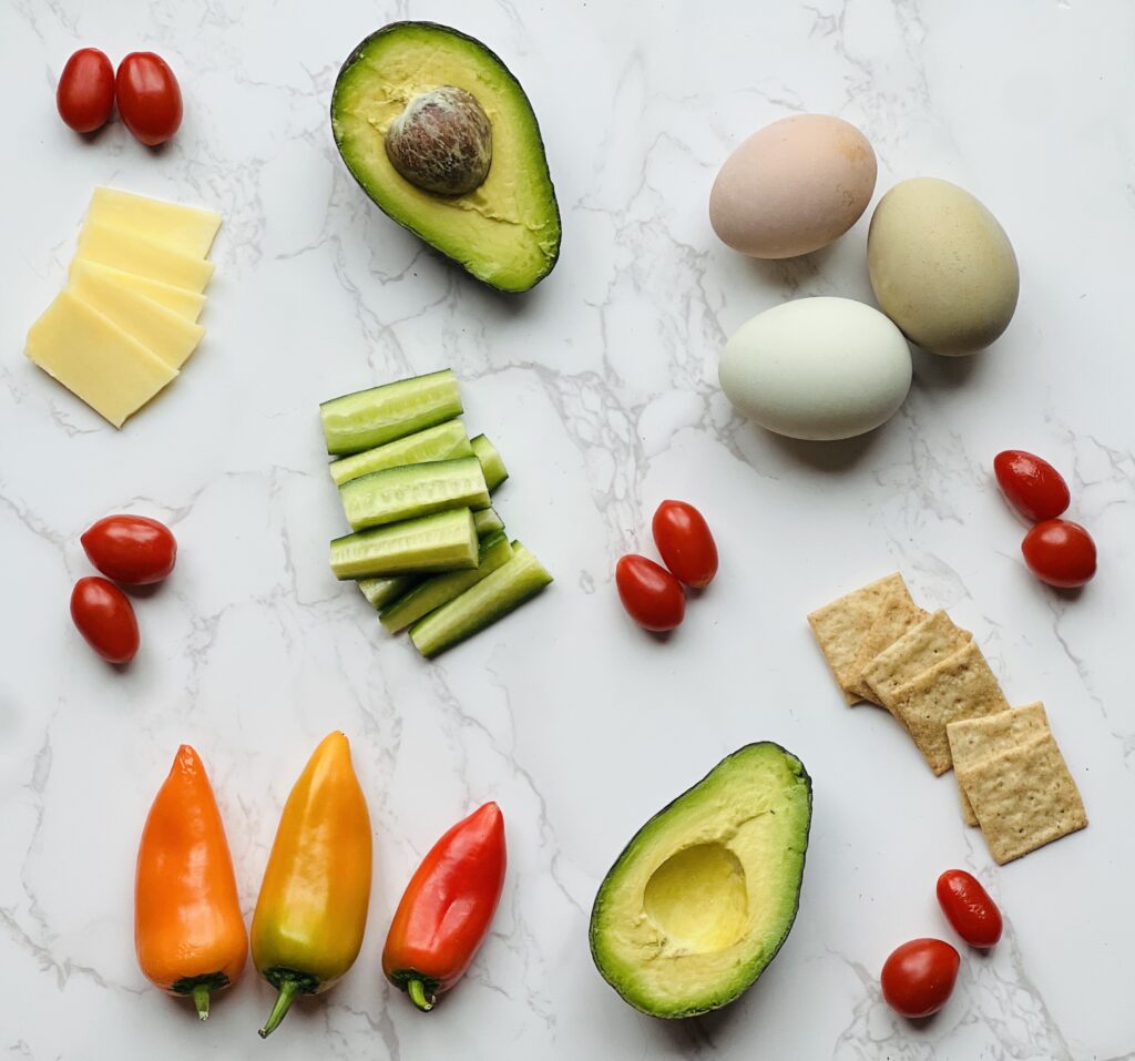 Some healthy whole food snacks such as hard boiled eggs, avocado, raw cheese, cucumbers, tomatoes, crackers and baby peppers lying on a marble countertop. 