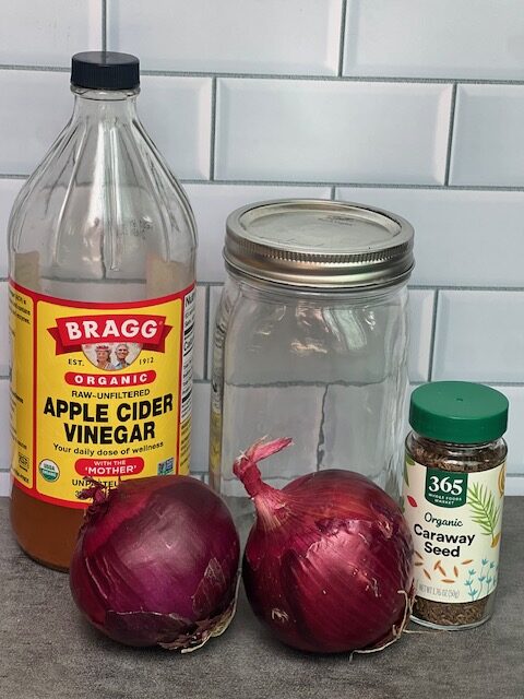 apple cider vinegar, a mason jar, 2 red onions uncut and a glass jar of caraway seeds sitting on a countertop