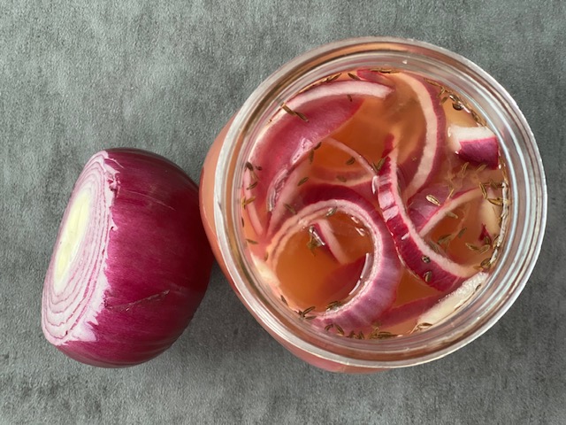 Freshly cut pickled red onions in a mason jar of apple cider vinegar and water with a half cut up red onion next to it
