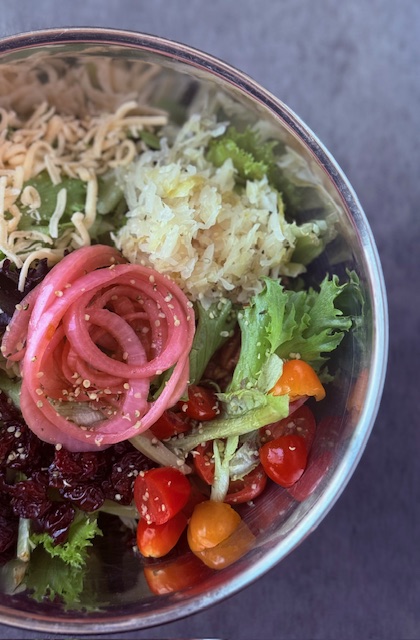 a beautifully colorful salad with cheese, cranberries, cherry tomatoes, sauerkraut and pickled onions atop it in a stainless steel bowl on a countertop