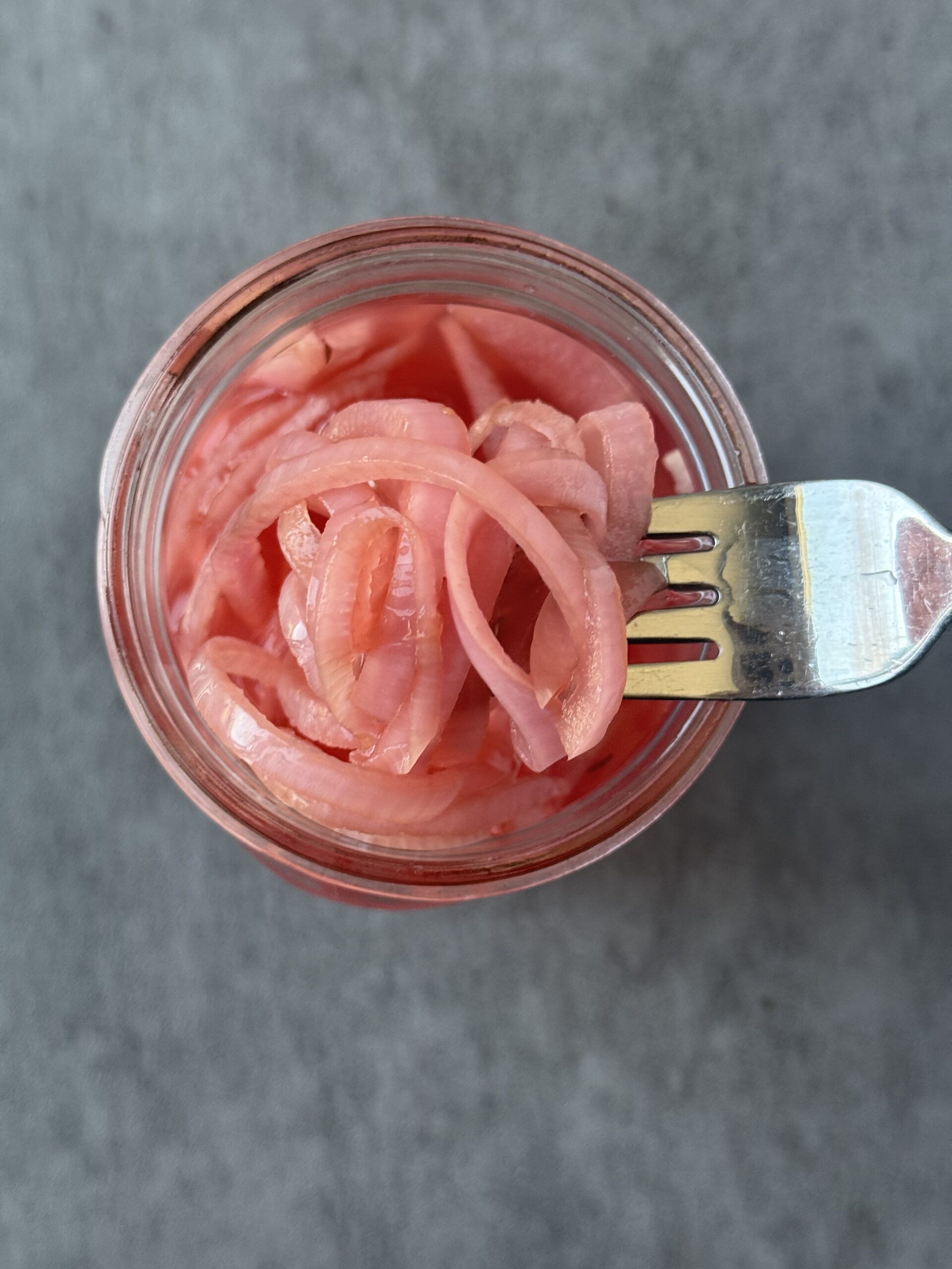 a silver fork scooping vibrant pink pickled red onion slices out of a mason jar full of an apple cider vinegar and water mixture.