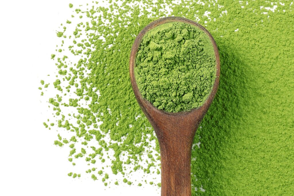Vibrant green matcha powder on a small wooden spoon with matcha powder all around it on a white countertop