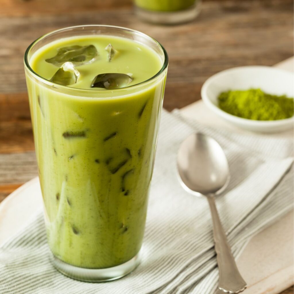 A glass of a green matcha latte with ice laying on a towel with a silver spoon next to it and a small of green matcha powder next to that. 