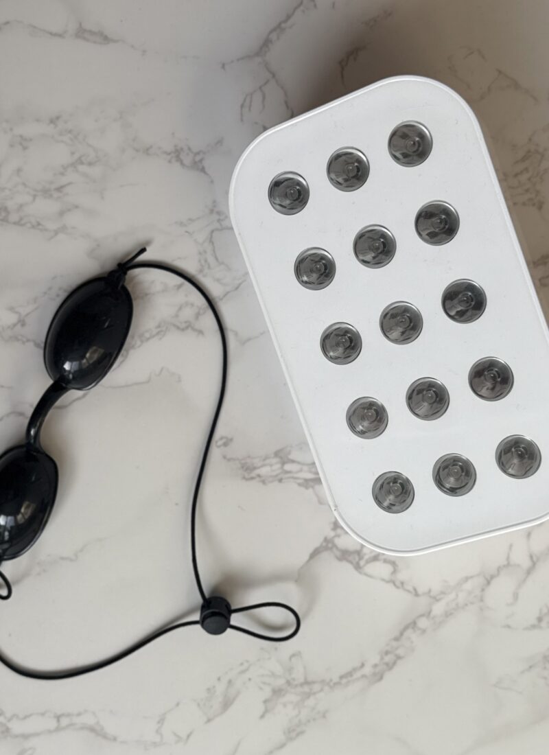 black eye goggles laid vertical on a white marble counter with a white red light therapy box next to it.