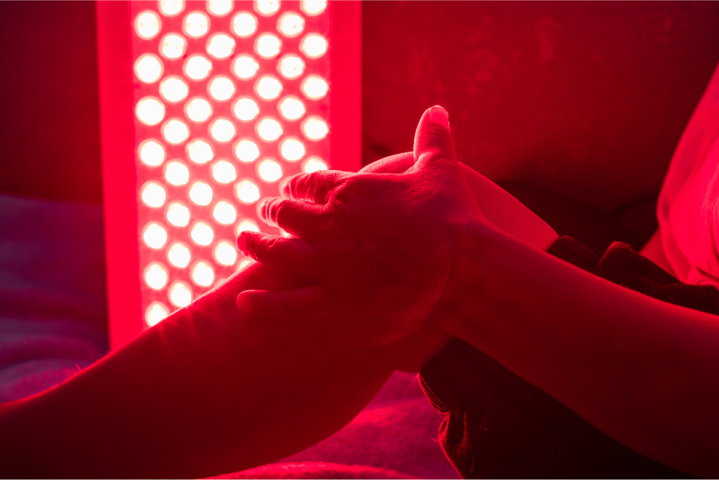a woman sitting on a bed with her knee up and her red light therapy box facing the knee and camera.
