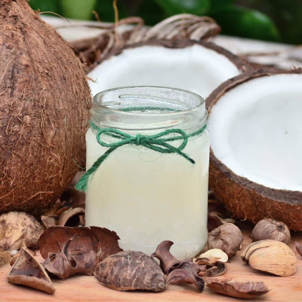 a jar of coconut oil sitting on a wood counter with 2 coconut halves and a whole coconut around it, with coconut shells on the counter near it.