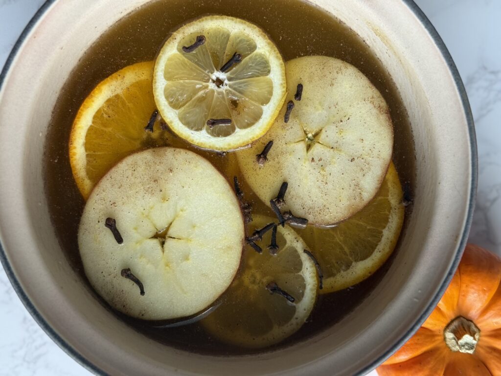 A pot with water and pumpkin puree mix along with slices of lemon, apple and orange, cinnamon sticks and cloves in it. It's sitting on a marble counter with a small orange pumpkin next to it.