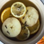 A pot with water and pumpkin puree mix along with slices of lemon, apple and orange, cinnamon sticks and cloves in it.