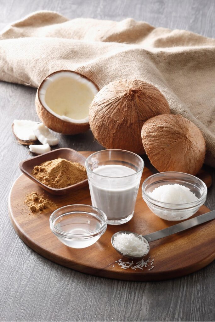 A wood tray on a floor with a glass of coconut milk, a bowl of liquid coconut oil, a bowl of solid coconut oil, a tablespoon of coconut flakes and a small tray of coconut sugar on it with a broken open coconut sitting next to them on the floor with a burlap blanket next to it all.