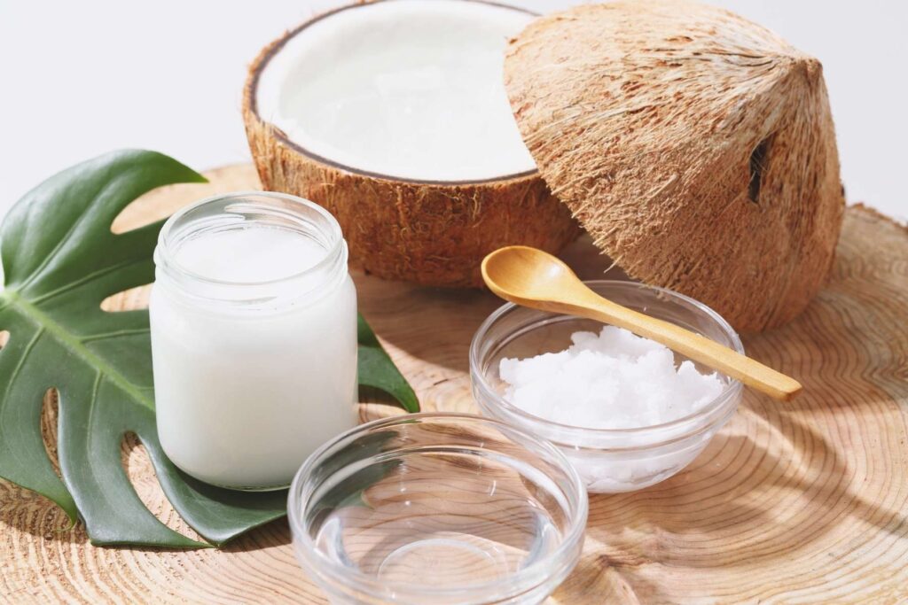 A jar of coconut oil, a bowl of coconut meat and a bowl of liquid coconut oil next to a cleanly sliced open coconut all sitting on a large tree stump.