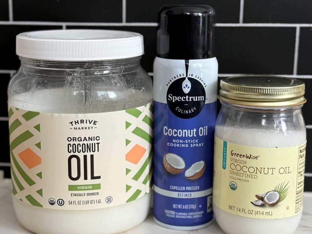 There is a container of coconut oil on the left next to a can of coconut oil cooking spray in the middle, with a glass jar of virgin unrefined coconut oil on the right all on a kitchen counter with a black tile background.