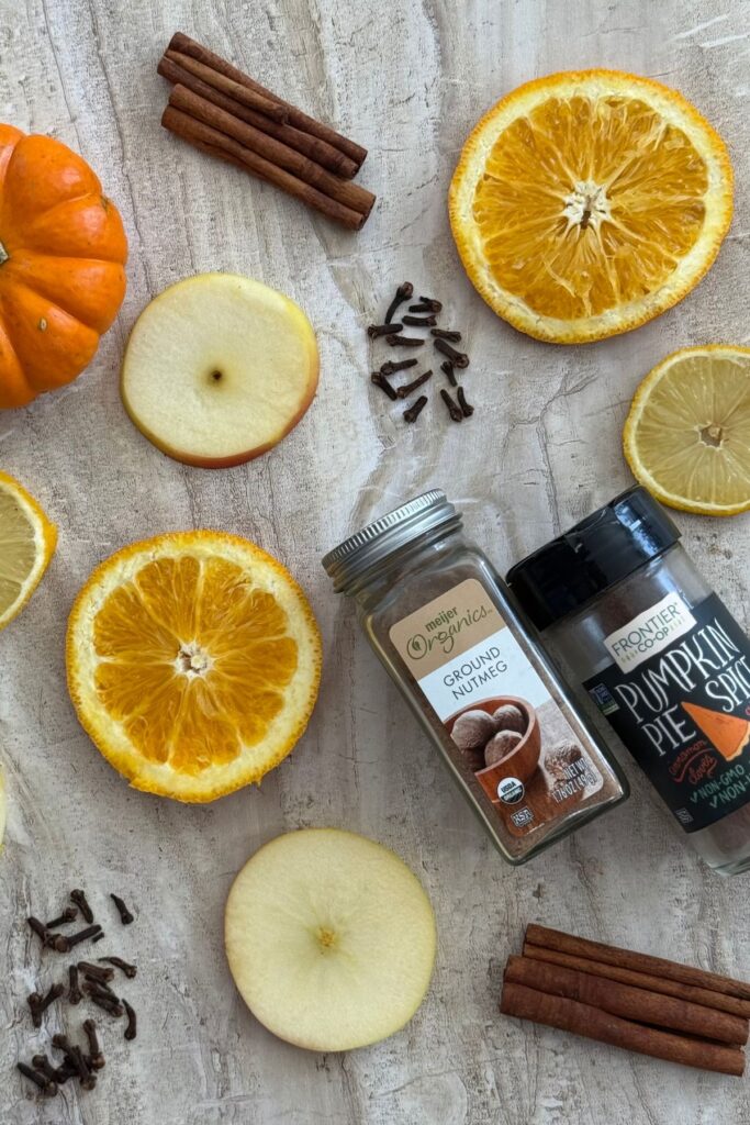 orange, apple and lemon slices on a counter with whole cloves, a few cinnamon sticks and a jar of nutmeg and a jar of pumpkin pie spice with a tiny pumpkin in the corner.