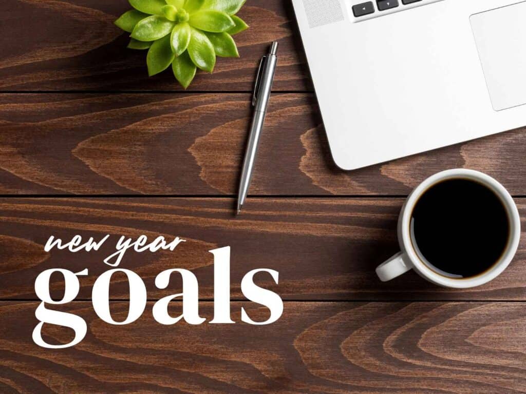 a wooden desk with a plant, laptop, pen and cup of coffee on it with the words: new year goals in the bottom left corner