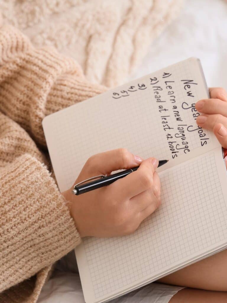 a woman's hands holding a journal that states New Years Goals and lists 1-6 with two goals written on the paper. 