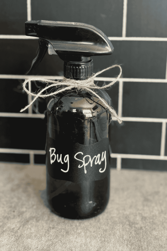 A homemade bug spray in a dark glass spray bottle with the words bug spray on it and a jute bow around the top