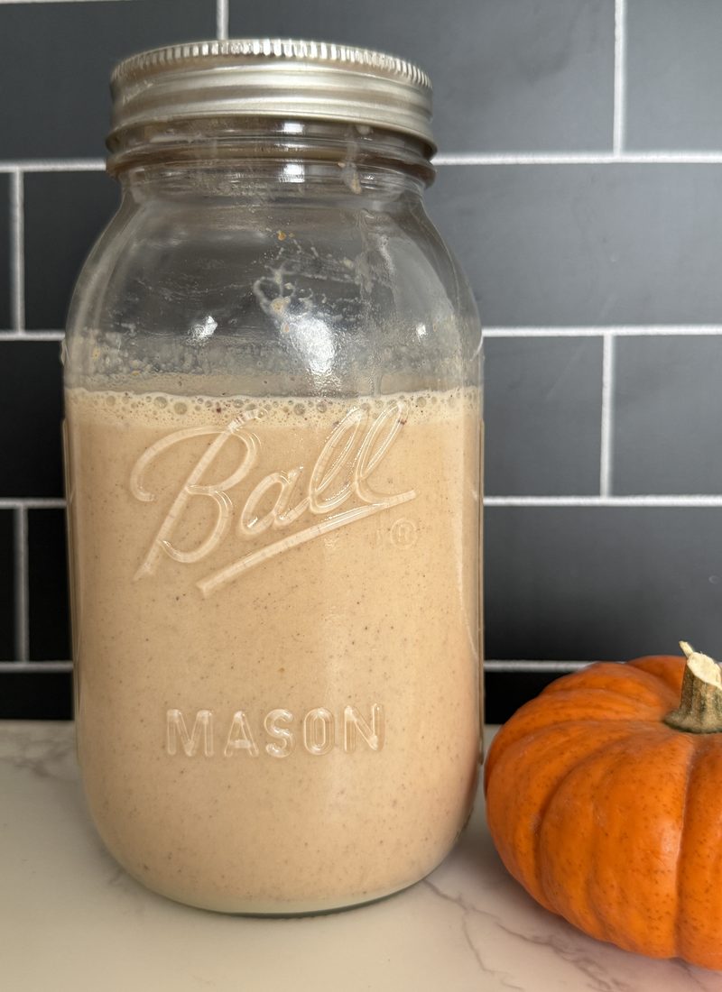 A mason jar full of a light brown/orange liquid that is a pumpkin spice coffee creamer. Next to the jar is a small orange pumpkin.