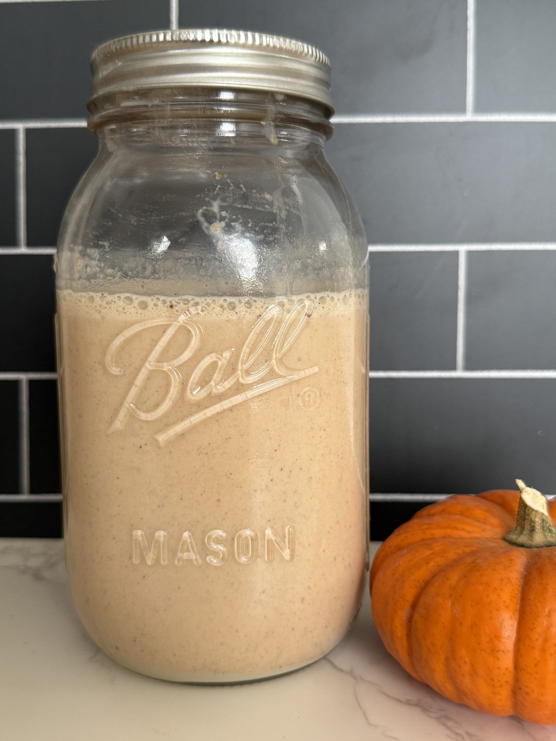 A mason jar full of a light brown/orange liquid that is a pumpkin spice coffee creamer. Next to the jar is a small orange pumpkin.