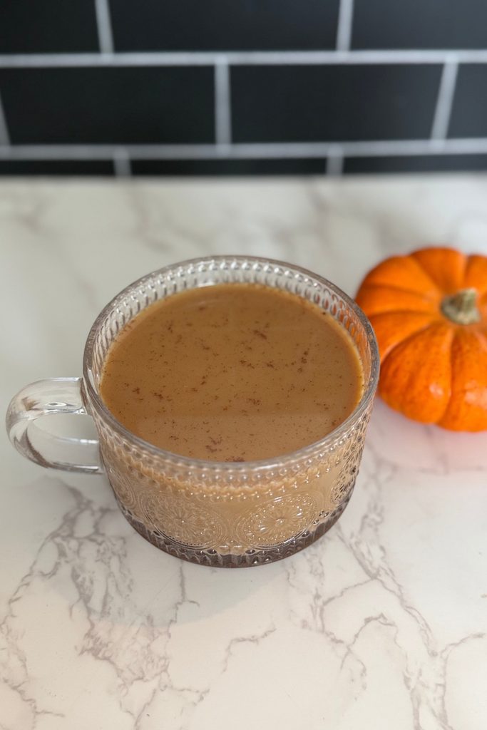 a cup of coffee with pumpkin spice creamer in it on a white counter top with black subway tile behind it and a small orange pumpkin to the back right corner of it.