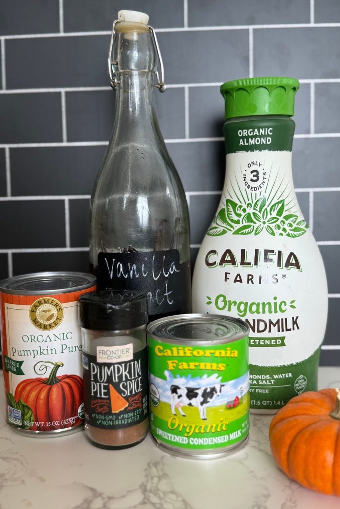 a white counter top with black subway tile background. On the counter are the ingredients needed to make a pumpkin spice creamer including almond milk, vanilla extract, pumpkin puree, pumpkin pie spice and sweetened condensed milk. There is a small pumpkin on the counter.